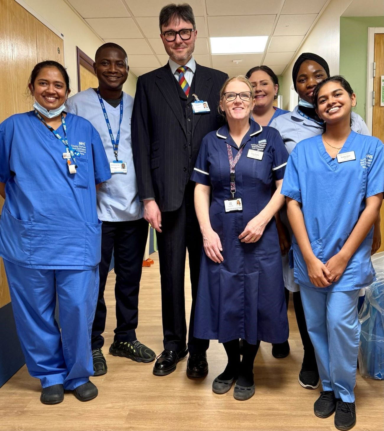 Man in suit stood with nurses in blue scrubs smiling on a ward