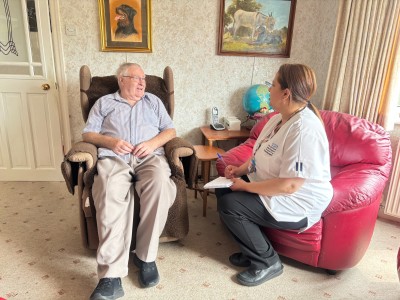 Man sat in chair at home with physiotherapist in white sat on sofa next to him